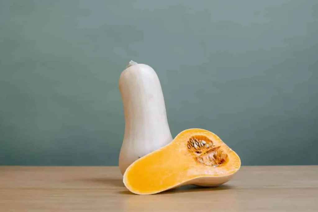 Courge butternut entière et coupée en deux, posée sur bois avec fond vert, révélant sa chair orange et ses graines.