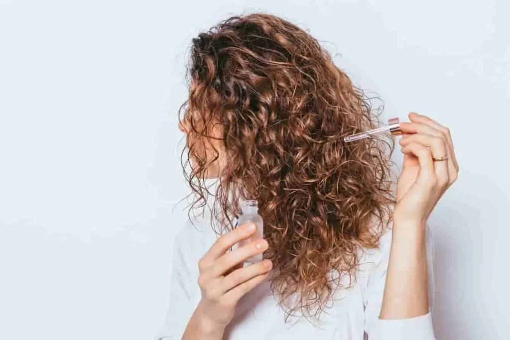 Personne en haut blanc versant de l’huile de coco sur ses boucles, fond épuré, geste de nutrition capillaire