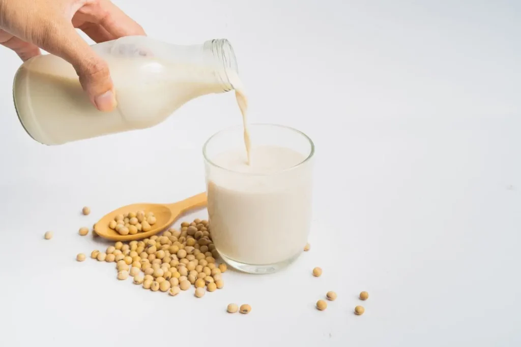 Lait de soja versé dans un verre, graines entières posées autour sur fond blanc, ambiance nutrition naturelle
