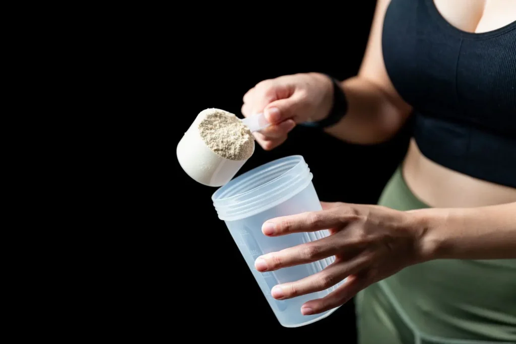 Personne en tenue sportive tenant une dosette de poudre beige au-dessus d’un shaker illustrant la préparation de créatine monohydrate pour musculation