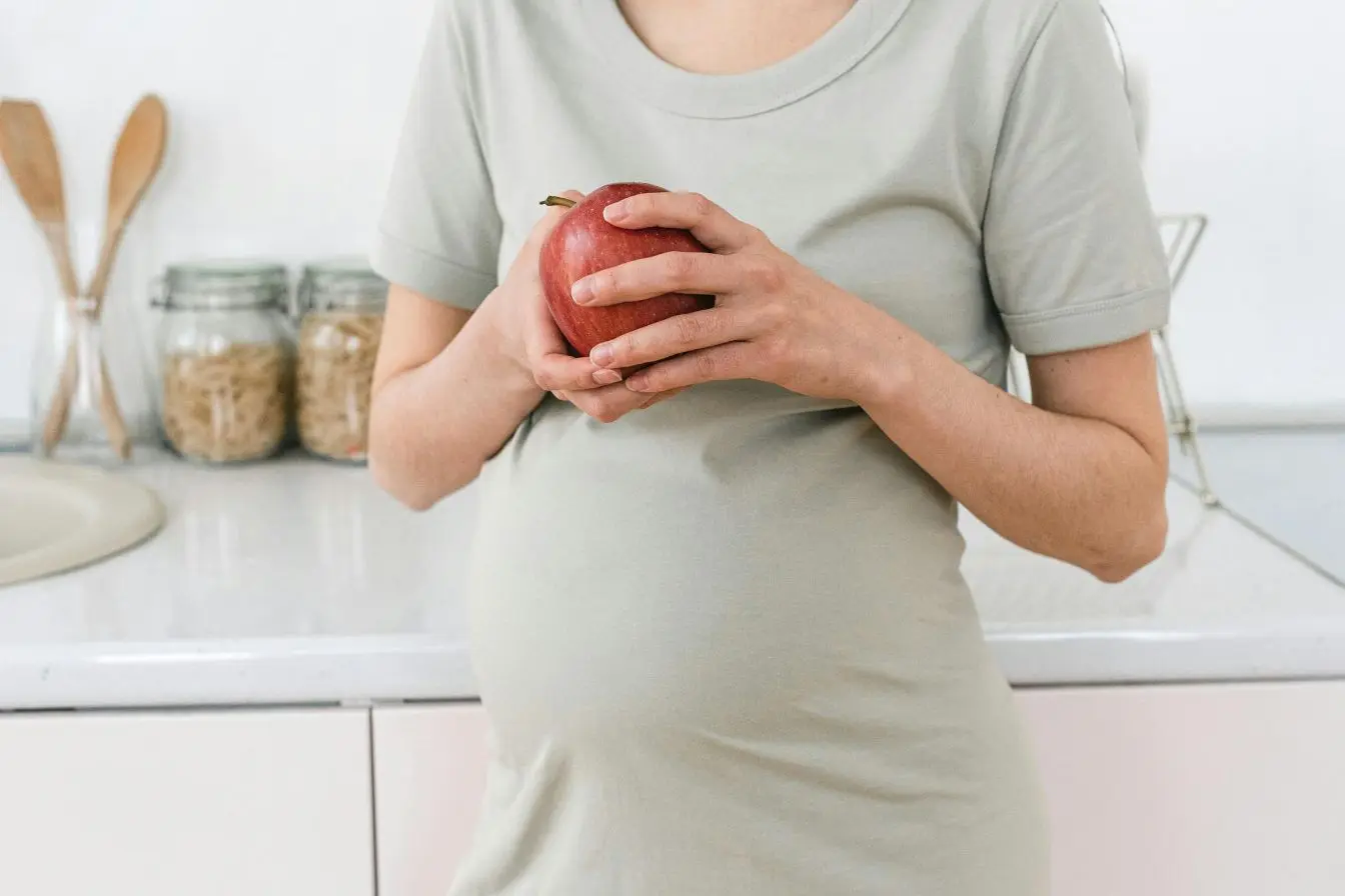 Femme enceinte tenant pomme rouge dans cuisine, image illustrant l’importance d’une alimentation saine au premier trimestre de grossesse.