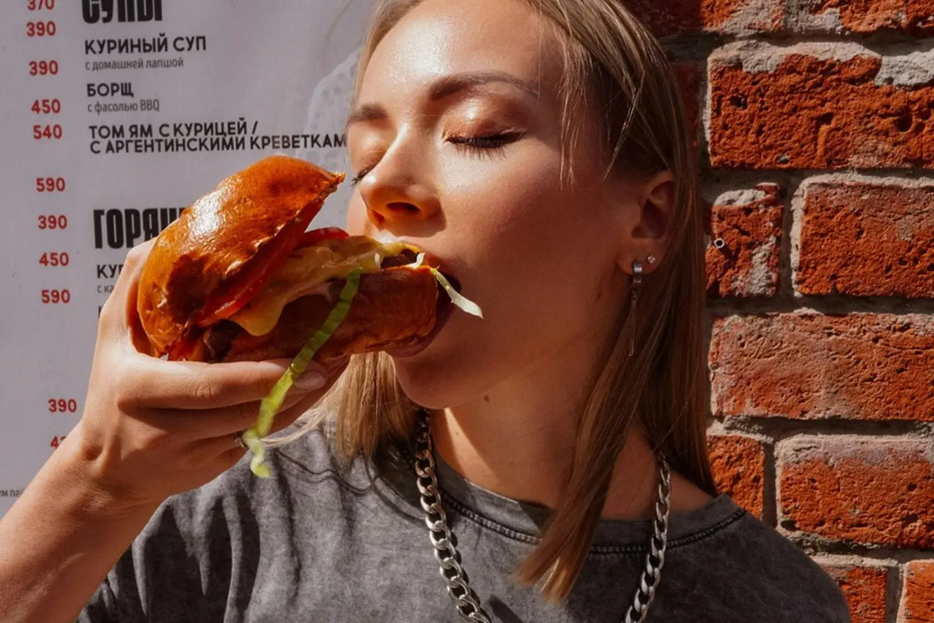 Personne mange un gros sandwich devant mur de briques, image illustrant la différence entre faim physique et appétit lié au désir gustatif.