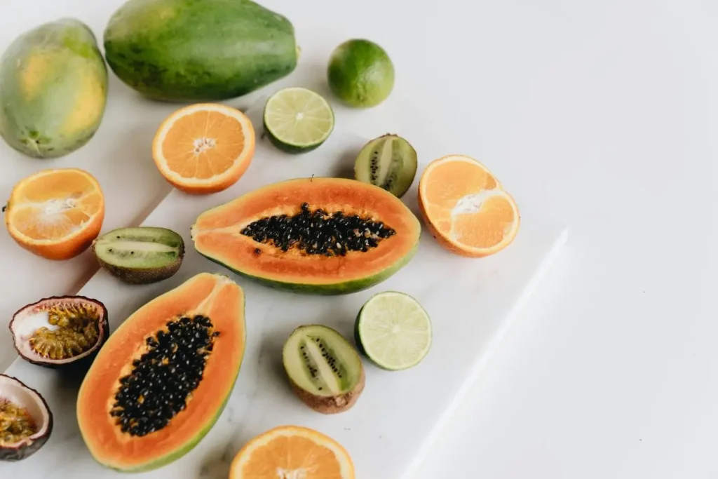 Papayes ouvertes avec chair orange et graines noires entourées d’oranges et kiwis, image illustrant les enzymes digestives naturelles des fruits tropicaux.