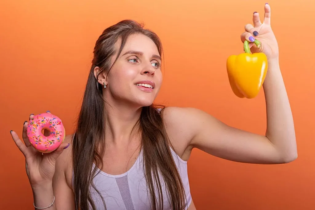 Femme tenant d’une main un donut rose et de l’autre un poivron jaune, image illustrant le lien entre alimentation et fertilité féminine.