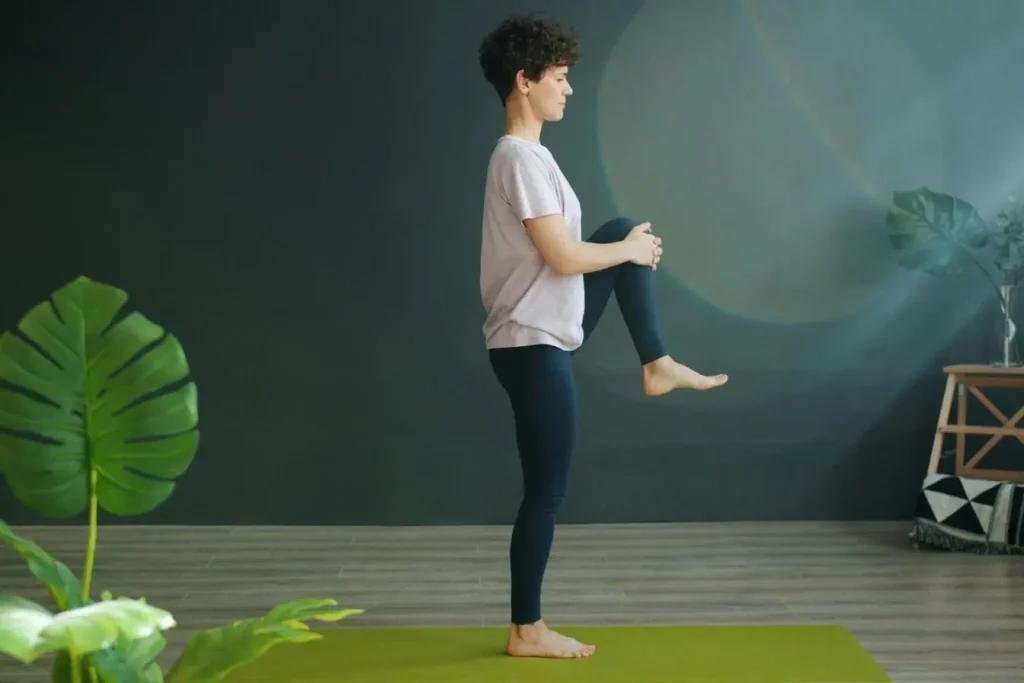 Personne debout sur tapis vert, genou levé et tenu par les mains, illustrant un exercice de proprioception et d’équilibre en intérieur.