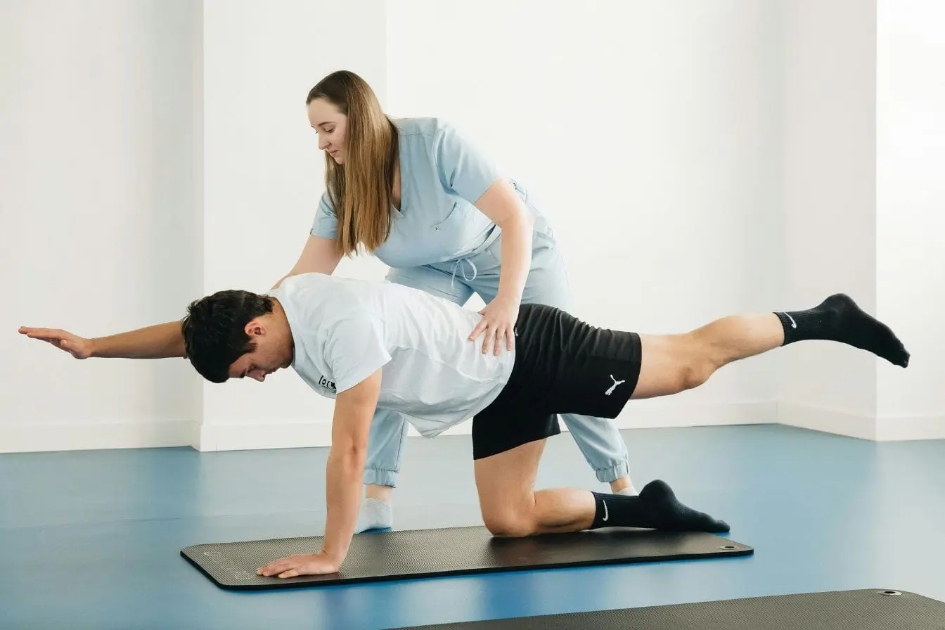 Patient en position quadrupède sur tapis, bras et jambe opposés étendus, guidé par kinésithérapeute pour exercices de proprioception et d’équilibre.
