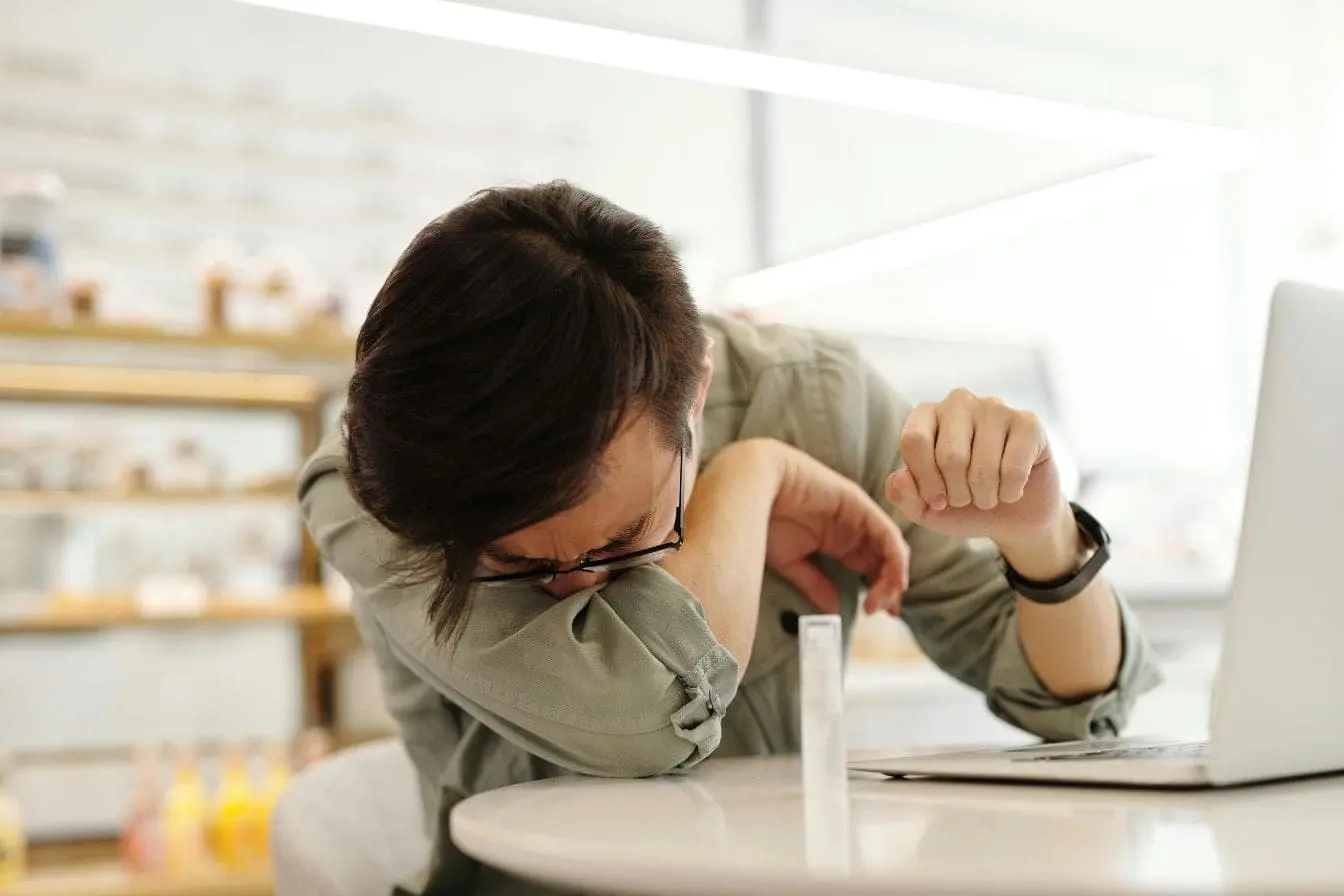 intolérance à l’histamine Personne assise à une table portant lunettes et chemise verte, se couvrant le visage en toussant, image illustrant l’intolérance à l’histamine.