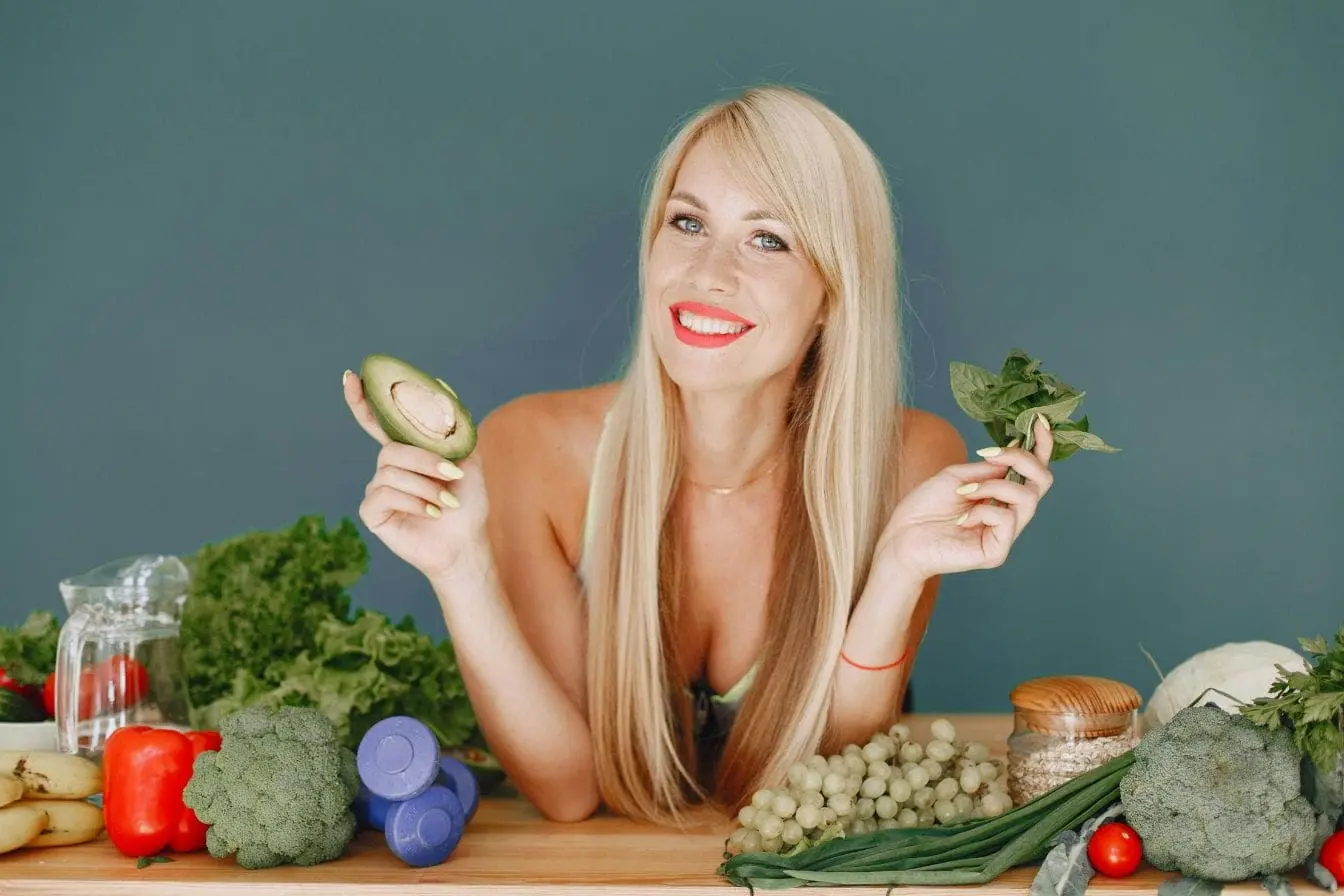 Personne souriante tenant avocat et légumes verts devant table remplie de fruits et graines, illustrant l’alimentation végane et santé équilibrée.