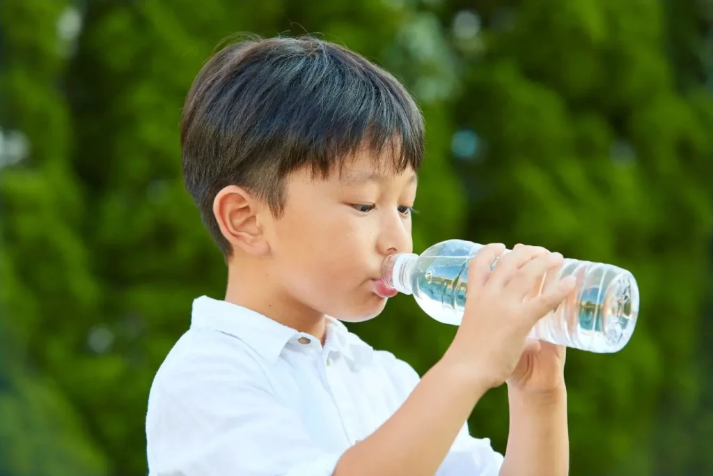 Enfant en chemise blanche boit eau d’une bouteille, image illustrant l’importance de l’hydratation dans l’alimentation des 3 à 6 ans.