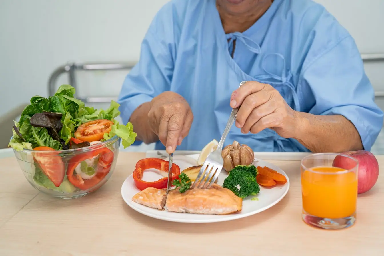 Plateau avec légumes variés Personne en blouse bleue déguste saumon et légumes colorés, image illustrant l’importance d’un repas équilibré pour répondre aux besoins nutritionnels des seniors.