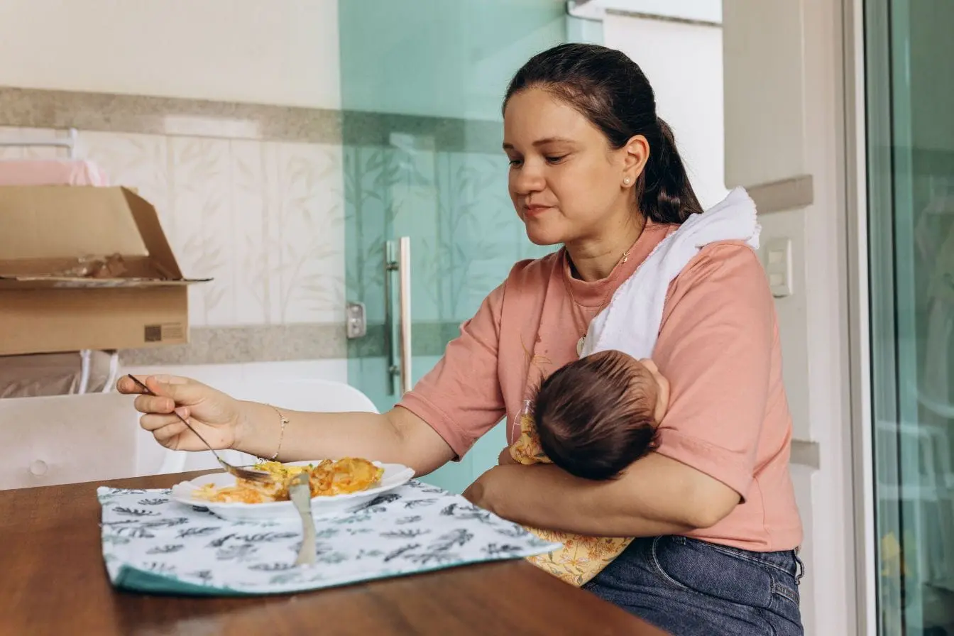 alimentation après accouchement Maman en chemise rose tient bébé emmailloté tout en mangeant, image illustrant l’importance de l’alimentation équilibrée après accouchement.