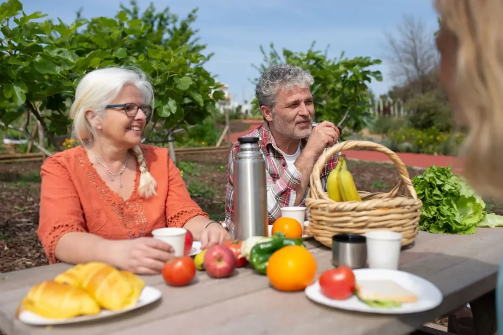 Deux seniors assis à table en plein air dégustent fruits et viennoiseries, image illustrant l’importance d’une alimentation équilibrée pour répondre aux besoins nutritionnels.
