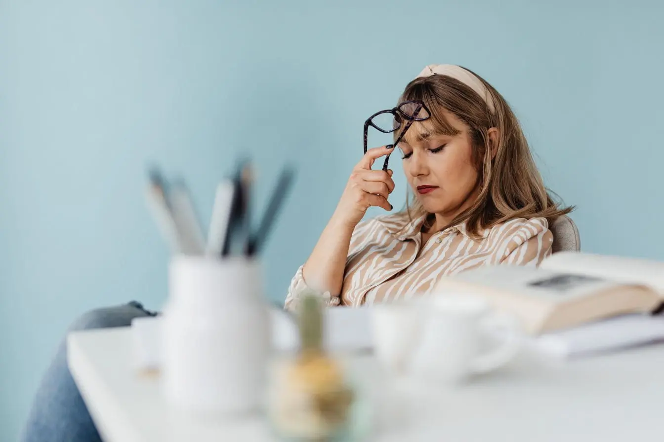 signes de surmenage Personne assise à un bureau, lunettes à la main, yeux fermés, exprimant la fatigue et les signes de surmenage liés au burn-out.