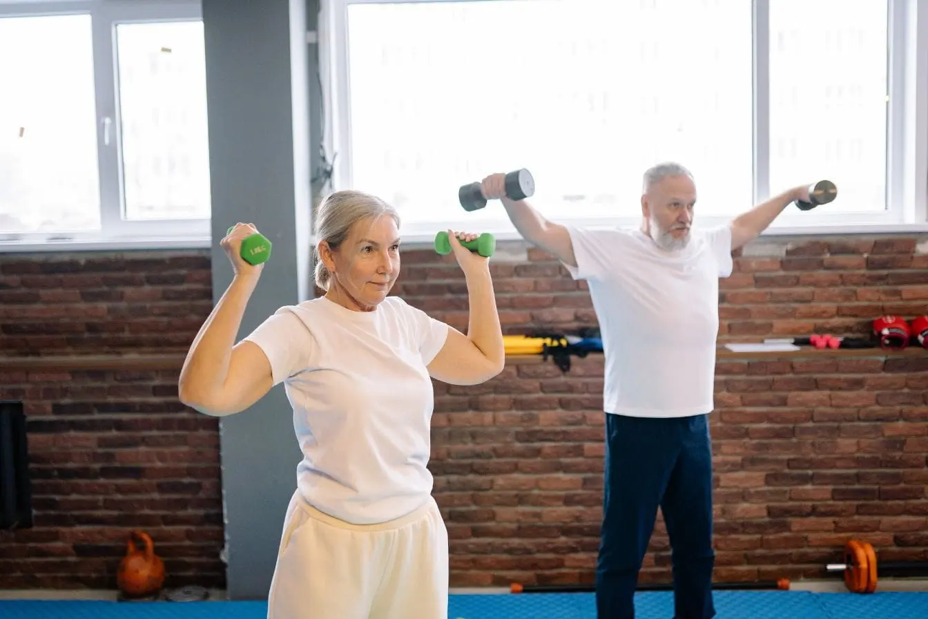 Deux personnes âgées en salle de sport tenant des haltères, image qui illustre l’importance du sport pour rester actif et en bonne santé.
