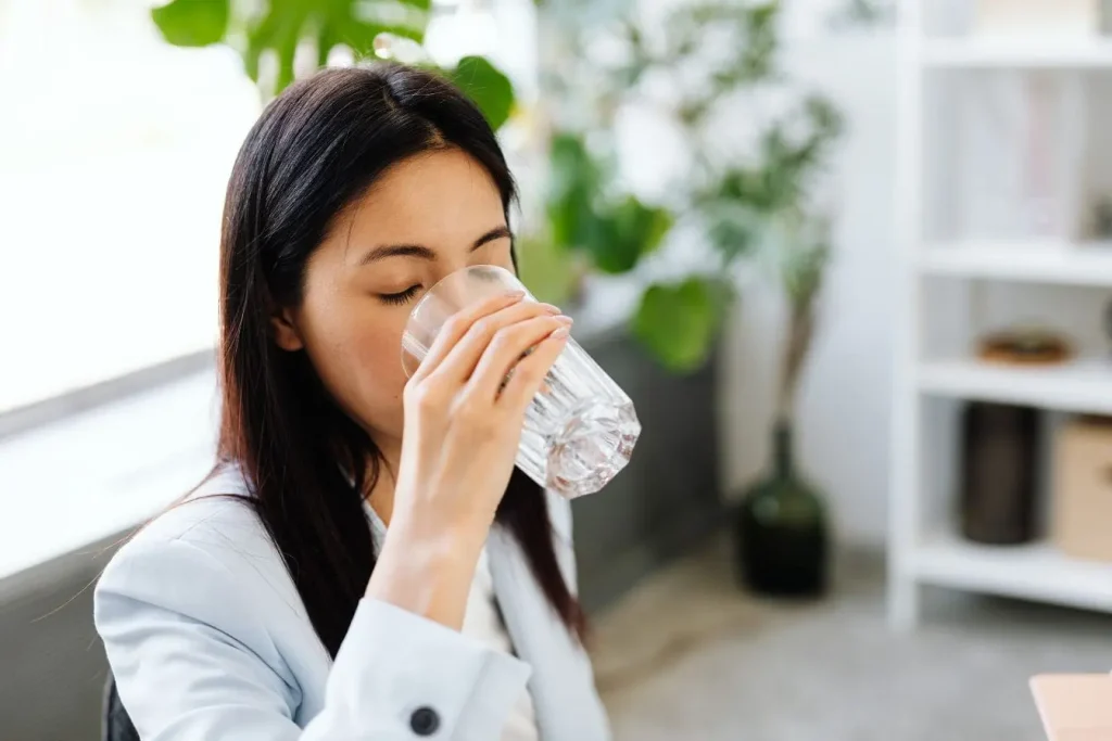 Personne assise dans intérieur lumineux buvant un verre d’eau, image qui illustre les bienfaits et méfaits de l’eau gazeuse pour la santé.