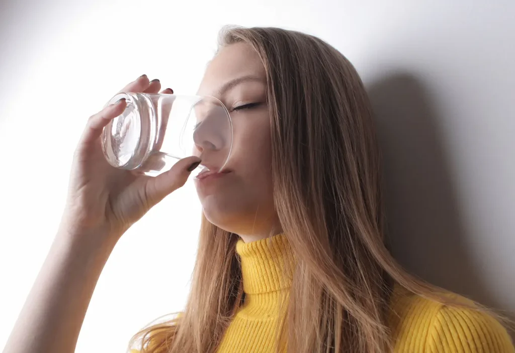 Personne en pull jaune buvant dans un verre transparent, geste reflétant les solutions naturelles contre la mauvaise haleine d’origine digestive.