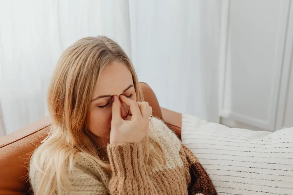 Une jeune femme assise sur un canapé se pince l'arête du nez avec fatigue, illustrant les céphalées et le stress liés à la spasmophilie.