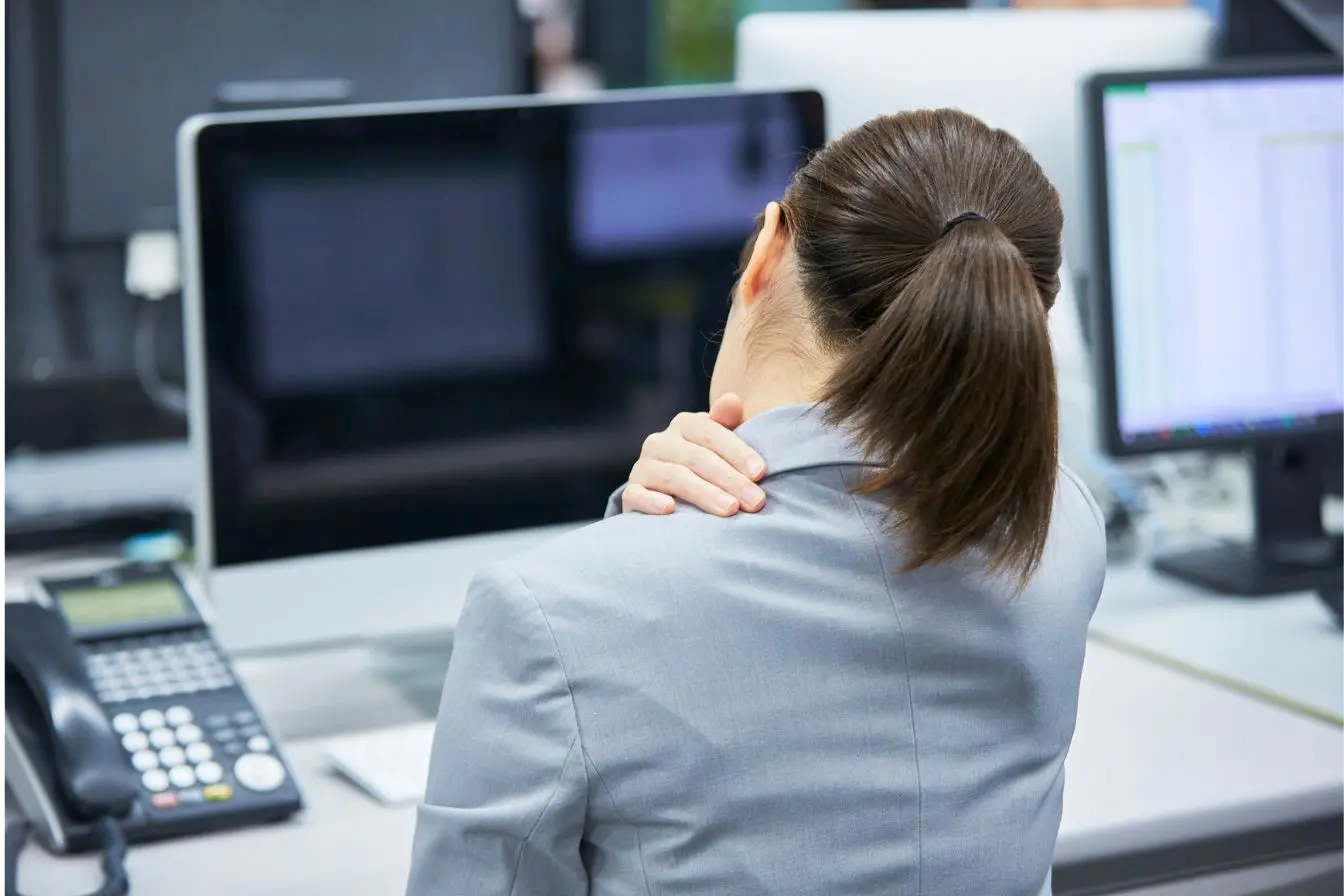 Torticolis Une employée de bureau assise devant son ordinateur se masse la nuque douloureuse pour trouver des remèdes rapides et gestes efficaces pour soulager le cou bloqué.