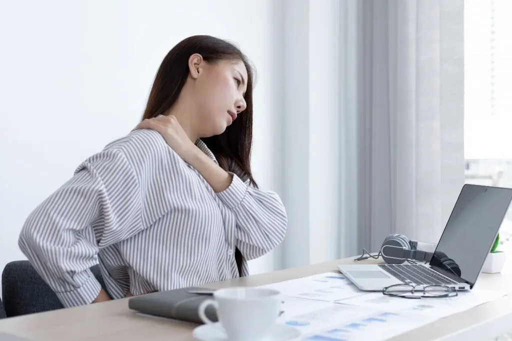 Une femme assise à son bureau devant un ordinateur portable qui se tient le dos et l'épaule en grimaçant, illustrant les douleurs liées au mal de dos chronique.