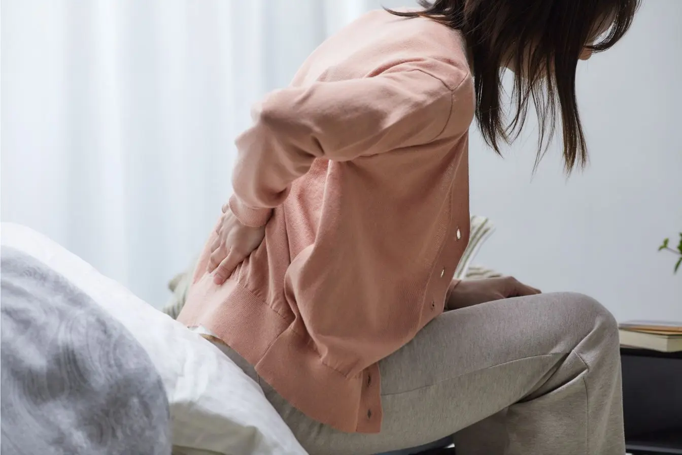 Une femme assise au bord de son lit tenant le bas de son dos avec une expression de douleur, illustrant le mal de dos chronique et les solutions naturelles.