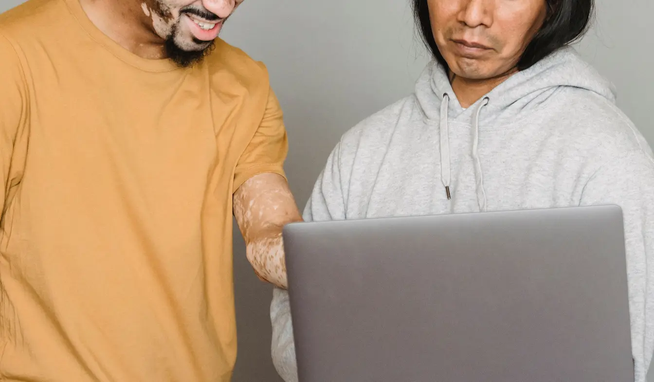Deux personnes debout regardant un ordinateur portable, l’une avec peau marquée, symbole des échanges autour du vitiligo et des solutions naturelles.